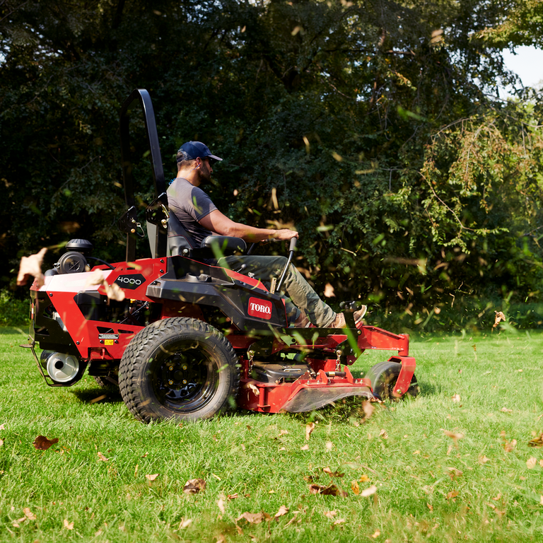 Man mowing a leaf-strewn lawn on a red Toro Z Master 4000 zero-turn mower in front of dense green trees