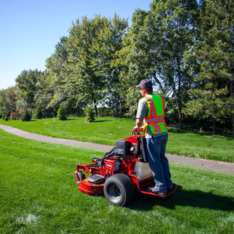 Landscaper in a safety vest mowing a park lawn on a red Toro GrandStand stand-on mower beside a curved path and tall trees