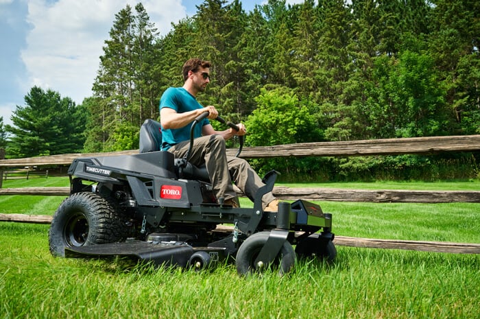 Man mowing a green lawn on a Toro TimeCutter zero-turn mower beside a split-rail fence and evergreen trees